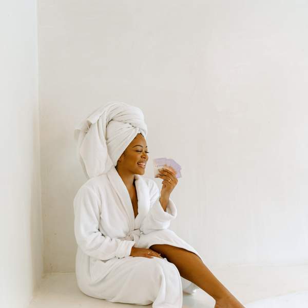 A Black woman in a white bathrobe with a white towel wrapped around her head smiles