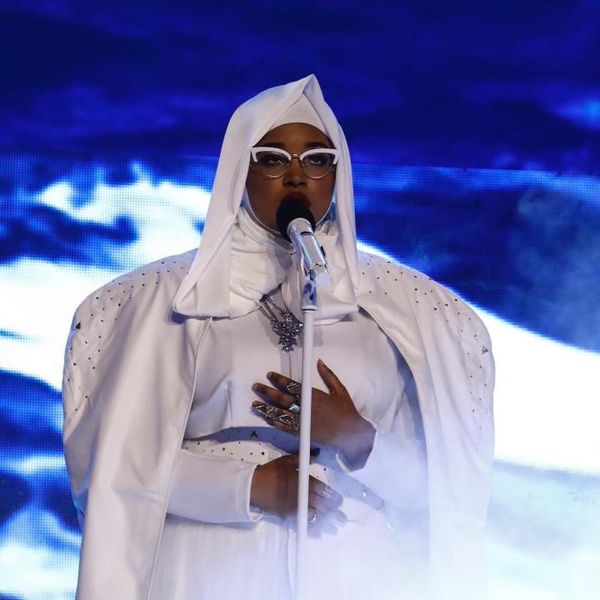 A Black woman in a white hijab, white cat-eyed glasses, white cape and outfit with rhinestones sings into a white microphone