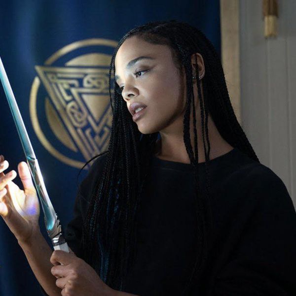A Black woman, Tessa Thompson, in character as Valkyrie, wearing long black box braids, and looking admiringly at the glowing sword she's holding