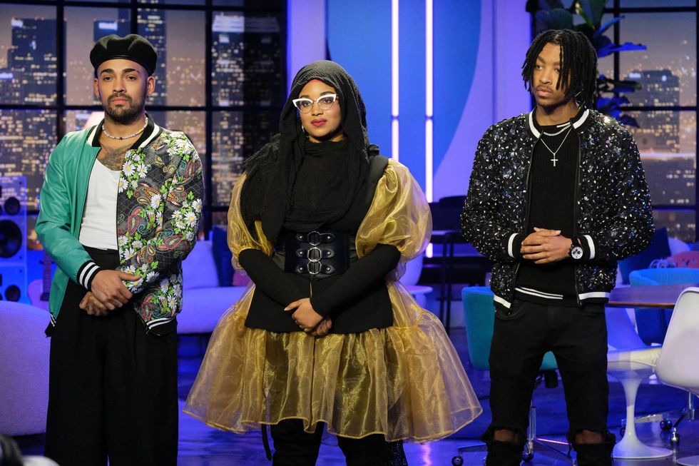 A Black woman wearing a black hijab and black and gold dress stands in between two men who are both wearing black pants and colorful jackets and necklaces