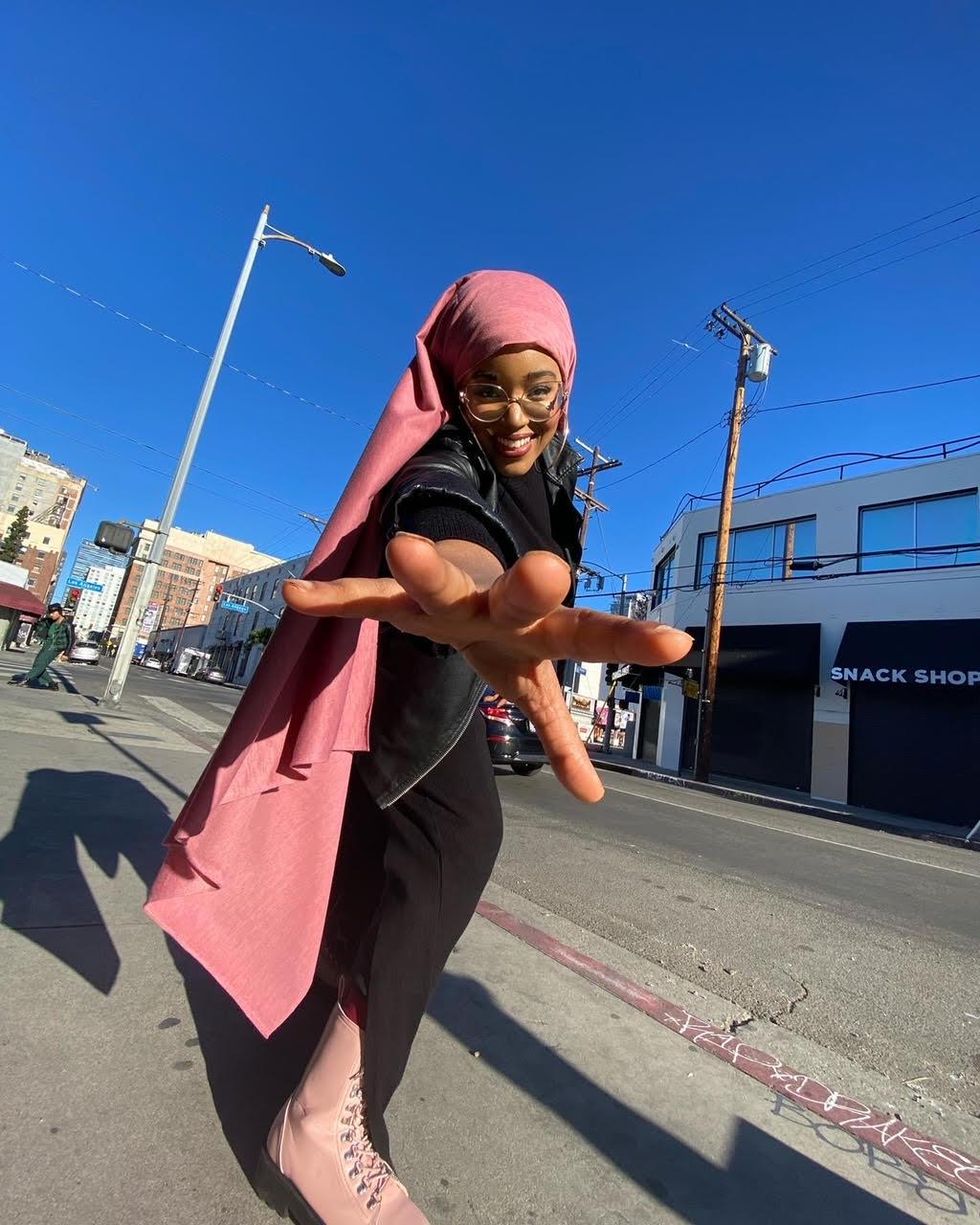 A Black woman wears a long, salmon pink hijab, black outfit and pink boots, smiling down at the camera with her arm outstretched to it.