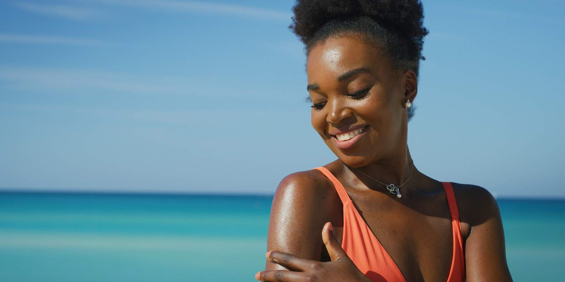 A happy young african woman is applying a sunscreen or sun tanning lotion to take care of her skin during a vacation on a beach.