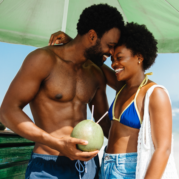 A loving couple shares a moment of affection during a baecation in Brazil. The tropical setting and clear skies highlight the joy and relaxation of their holiday, as they hold a coconut drink.