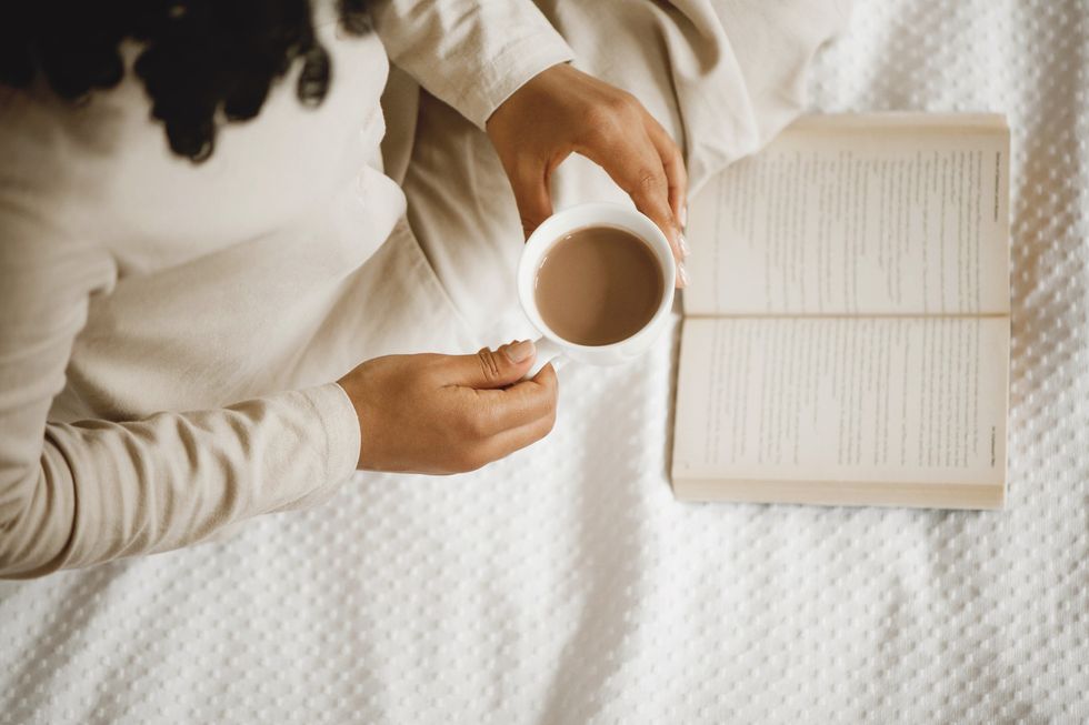 aerial-view-of-a-woman-in-beige-pajamas-holding-a-warm-beverage-reading-a-book-against-a-white-blanket