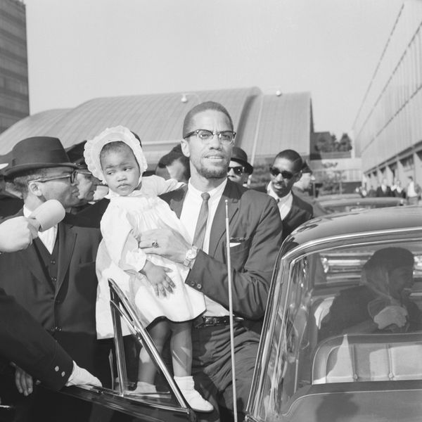 black and white image of Malcolm X in a suit, tie and glasses, stepping into a car while holding his toddler daughter who's wearing a bonnet, dress and dress shoes and shocks.