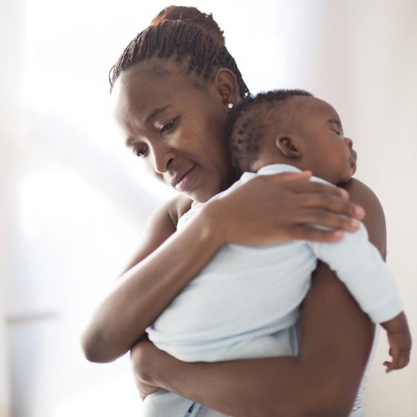 Black mother with brown microbraids and a diamond earring in her ear looks sad as she holds a sleeping infant wearing a blue onesie