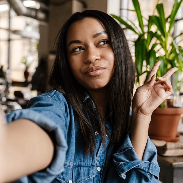 Black smiling woman gesturing peace sign and taking selfie photo while sitting in cafe