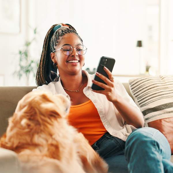 Black woman relaxing on sofa with dog while scrolling on smartphone