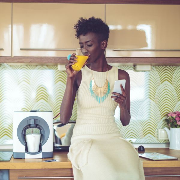 Black-woman-sitting-on-her-kitchen-counter-drinking-juice-while-holding-her-phone