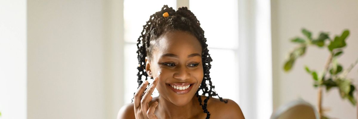 Cheerful black lady wrapped in towel using facial cream after shower, making skin ready for makeup, sitting at table at home, looking at mirror, free space