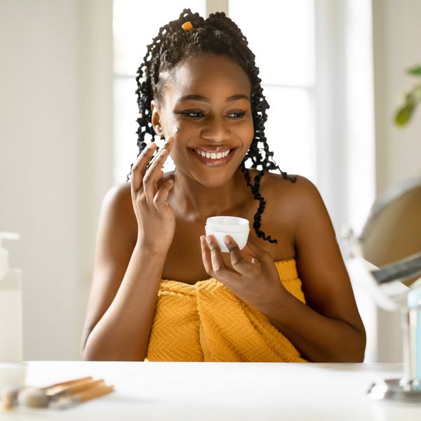 Cheerful black lady wrapped in towel using facial cream after shower, making skin ready for makeup, sitting at table at home, looking at mirror, free space