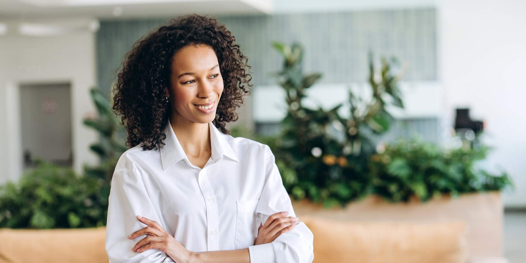 Confident businesswoman is crossing her arms and smiling while standing in a modern office with plants and comfortable furniture, showcasing leadership and professionalism looking away, copy space