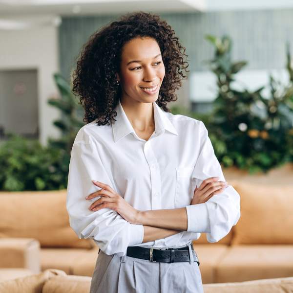 Confident businesswoman is crossing her arms and smiling while standing in a modern office with plants and comfortable furniture, showcasing leadership and professionalism looking away, copy space