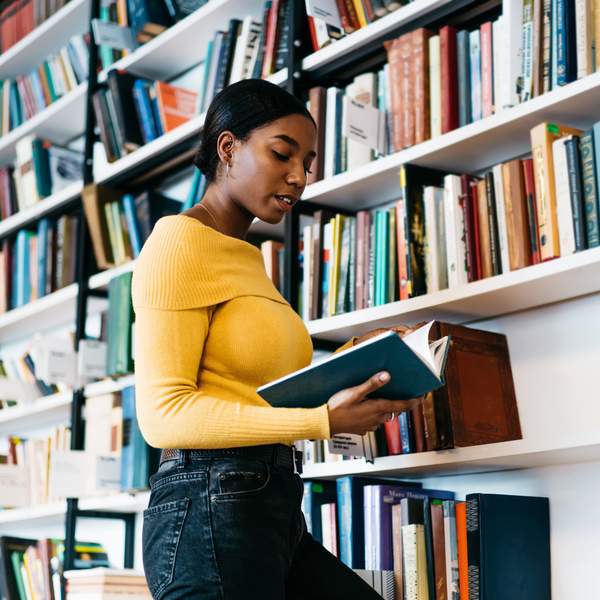 Focused African American lady in casual clothes reading textbook while standing near bookcase and studying in library of contemporary university