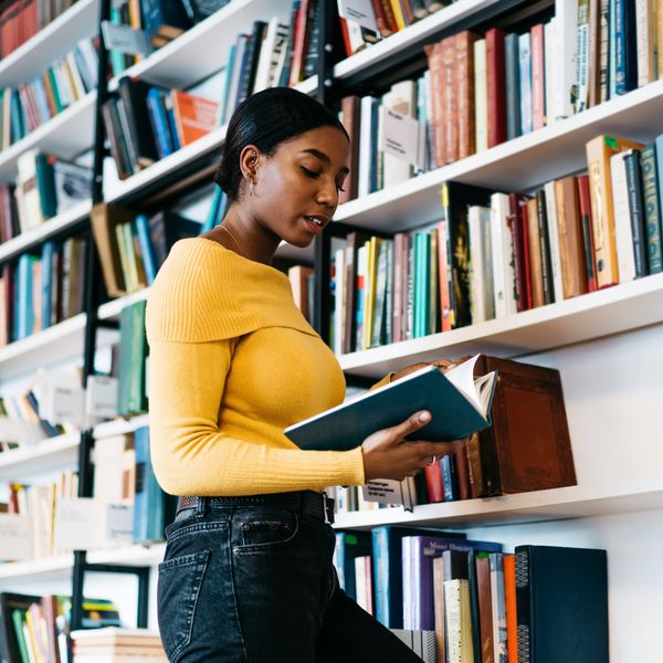 Focused African American lady in casual clothes reading textbook while standing near bookcase and studying in library of contemporary university