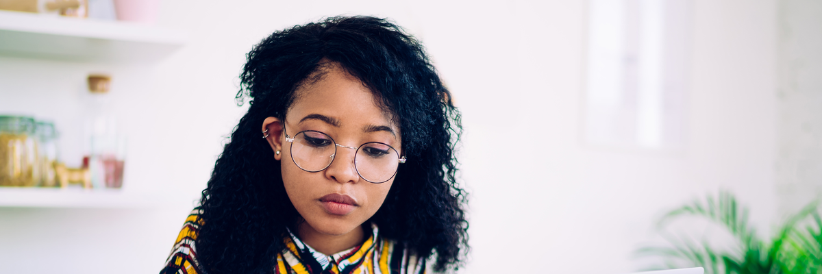 Focused ethnic female freelancer in glasses browsing smartphone and laptop while sitting at table with cup of coffee at home