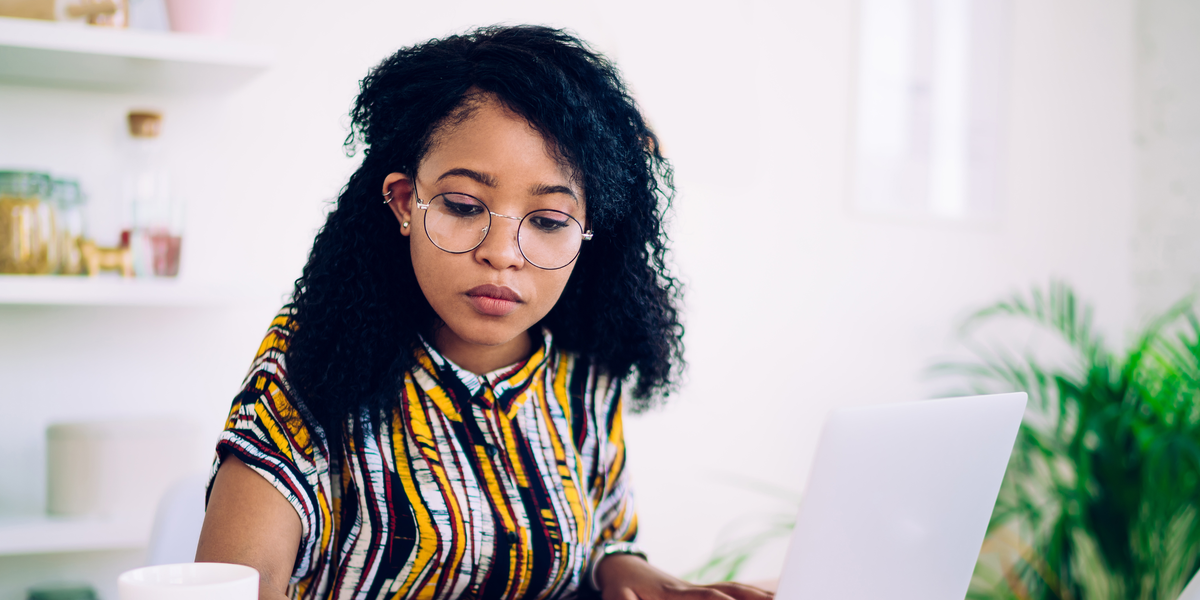 Focused ethnic female freelancer in glasses browsing smartphone and laptop while sitting at table with cup of coffee at home