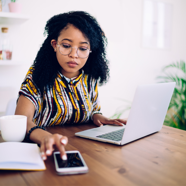 Focused ethnic female freelancer in glasses browsing smartphone and laptop while sitting at table with cup of coffee at home