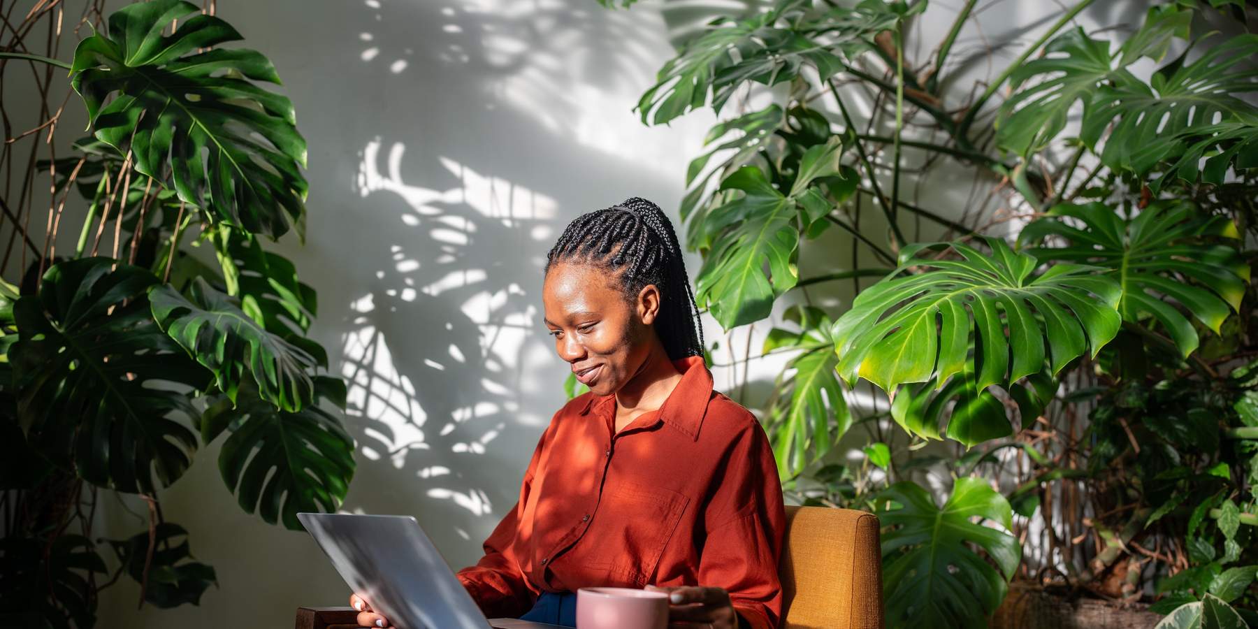Giggling black woman using laptop. Cheerful laughing african american girl in chair among plants, drinking coffee from mug looking at computer screen, watching video movie series online on internet.