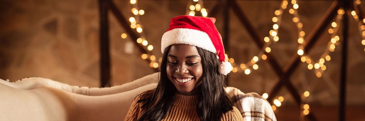 Happy black lady in Santa hat using laptop, ordering Xmas gifts on web, shopping for Christmas presents online from home. African American woman sitting on couch with computer on winter holidays