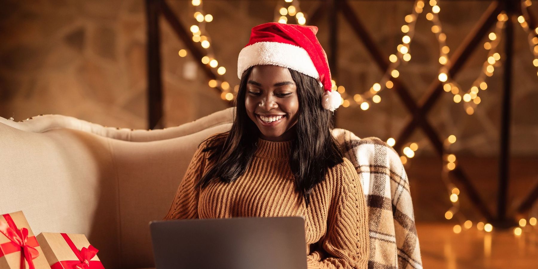 Happy black lady in Santa hat using laptop, ordering Xmas gifts on web, shopping for Christmas presents online from home. African American woman sitting on couch with computer on winter holidays