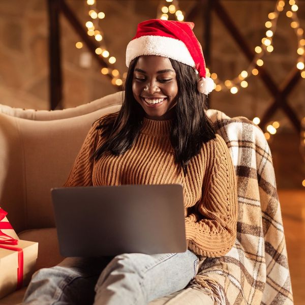Happy black lady in Santa hat using laptop, ordering Xmas gifts on web, shopping for Christmas presents online from home. African American woman sitting on couch with computer on winter holidays