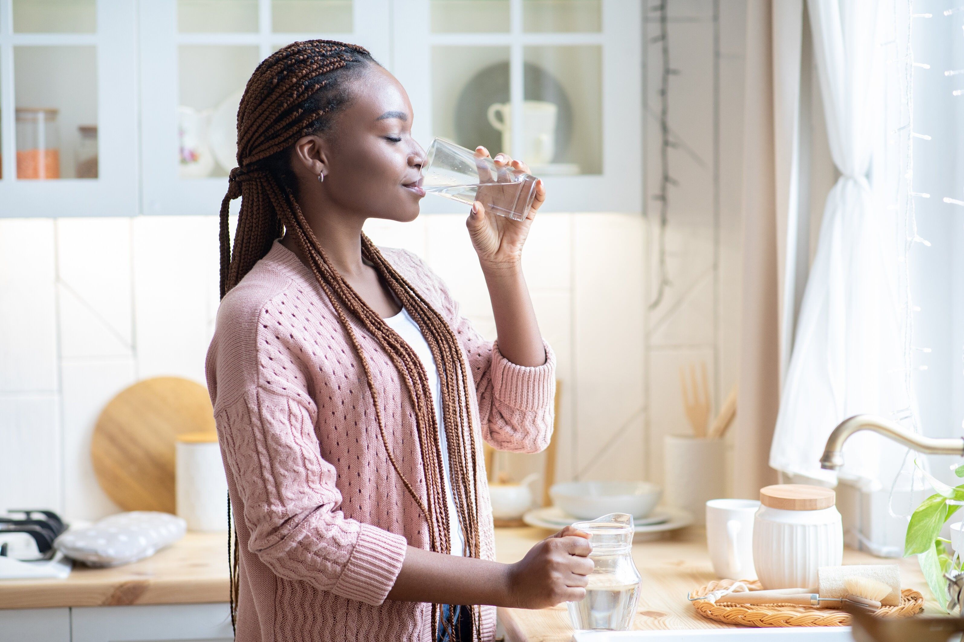Thirsty African American Woman Drinking Water From Glass In Kitchen At Home, Young Black Female Standing Near Counter And Enjoying Heathy Refreshing Drink, Side View With Copy Space