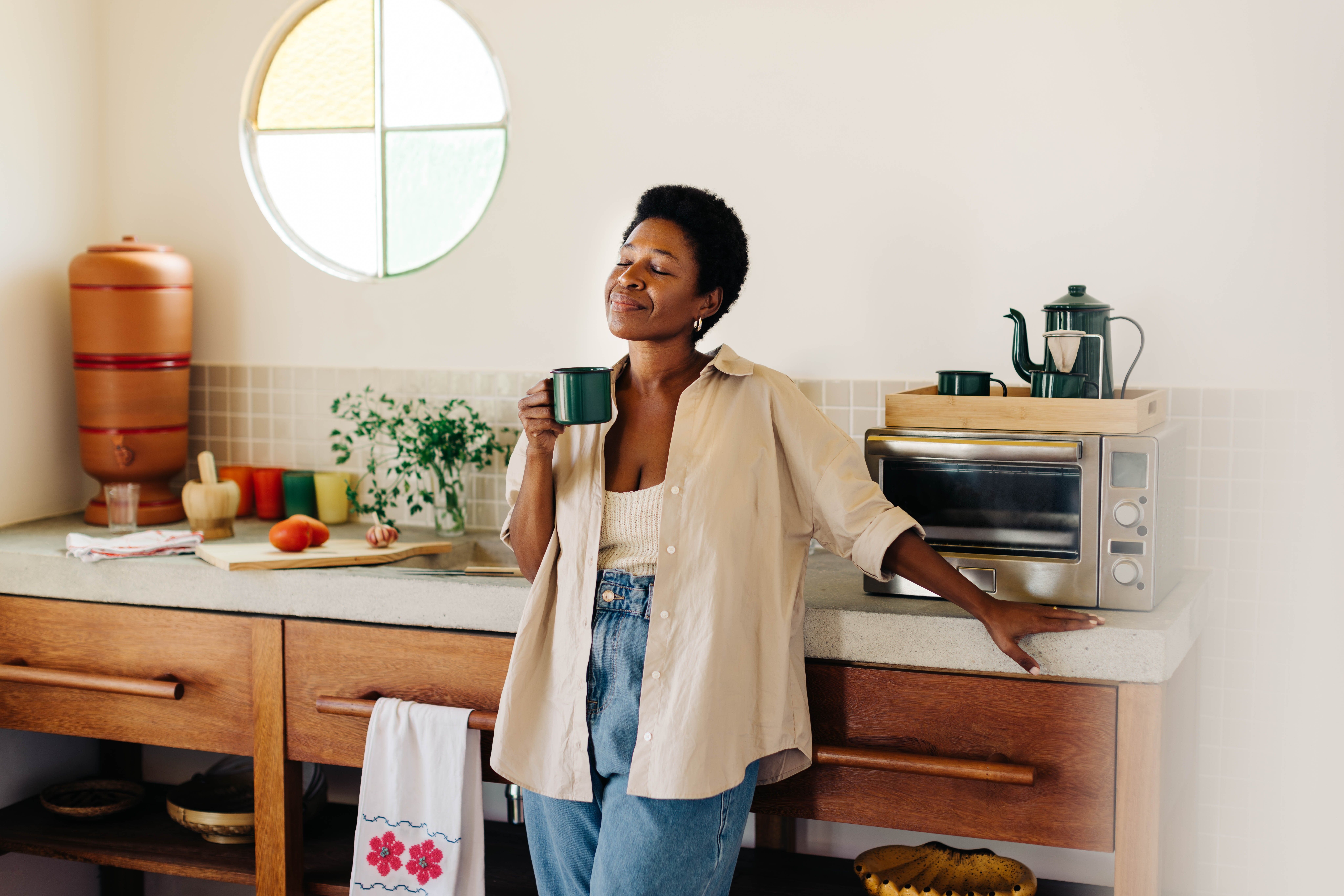 Happy Brazilian woman with afro hair stands in her kitchen, holding an enamel mug of hot coffee, eyes closed, enjoying a peace and quiet moment at home.
