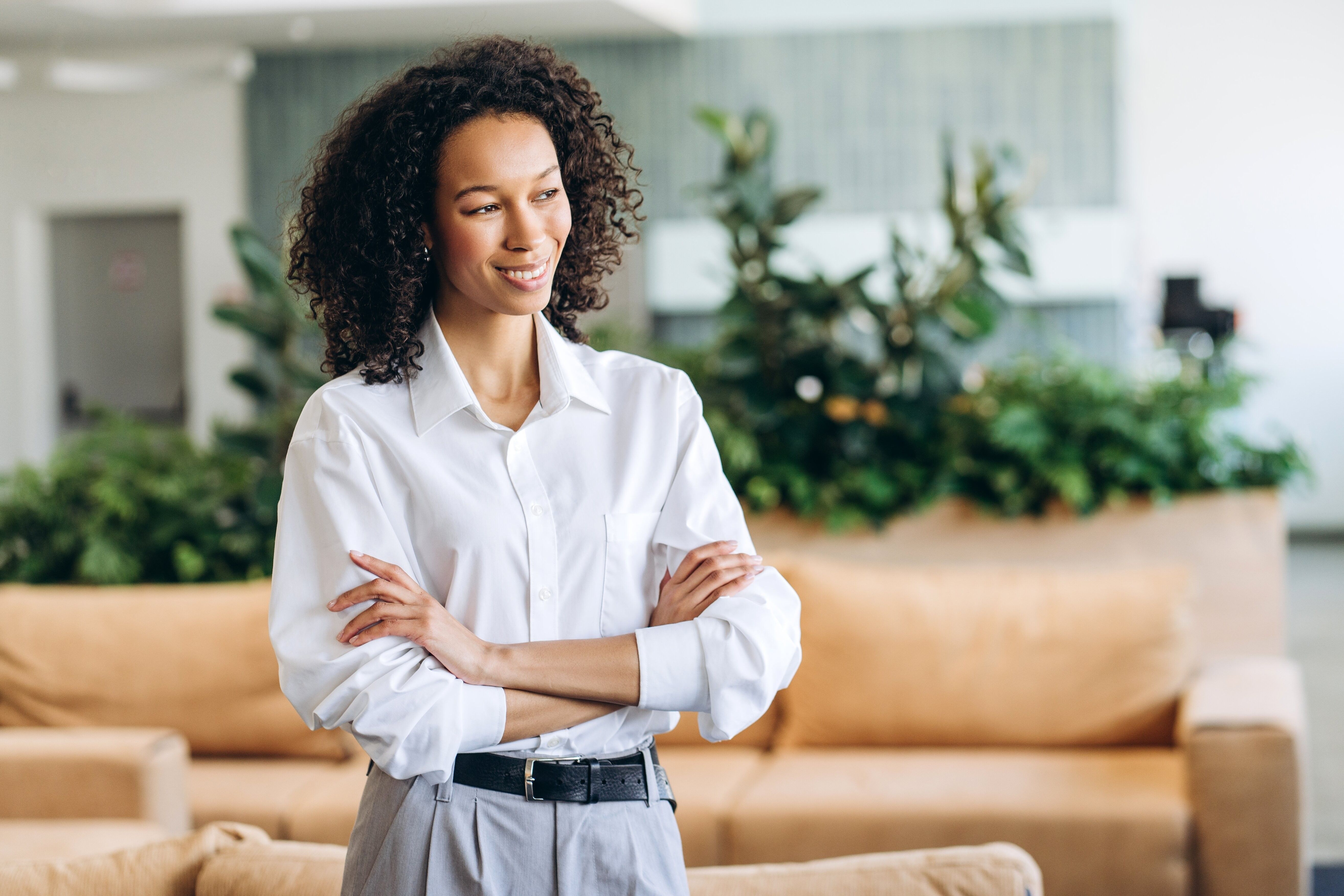 Confident businesswoman is crossing her arms and smiling while standing in a modern office with plants and comfortable furniture, showcasing leadership and professionalism looking away, copy space