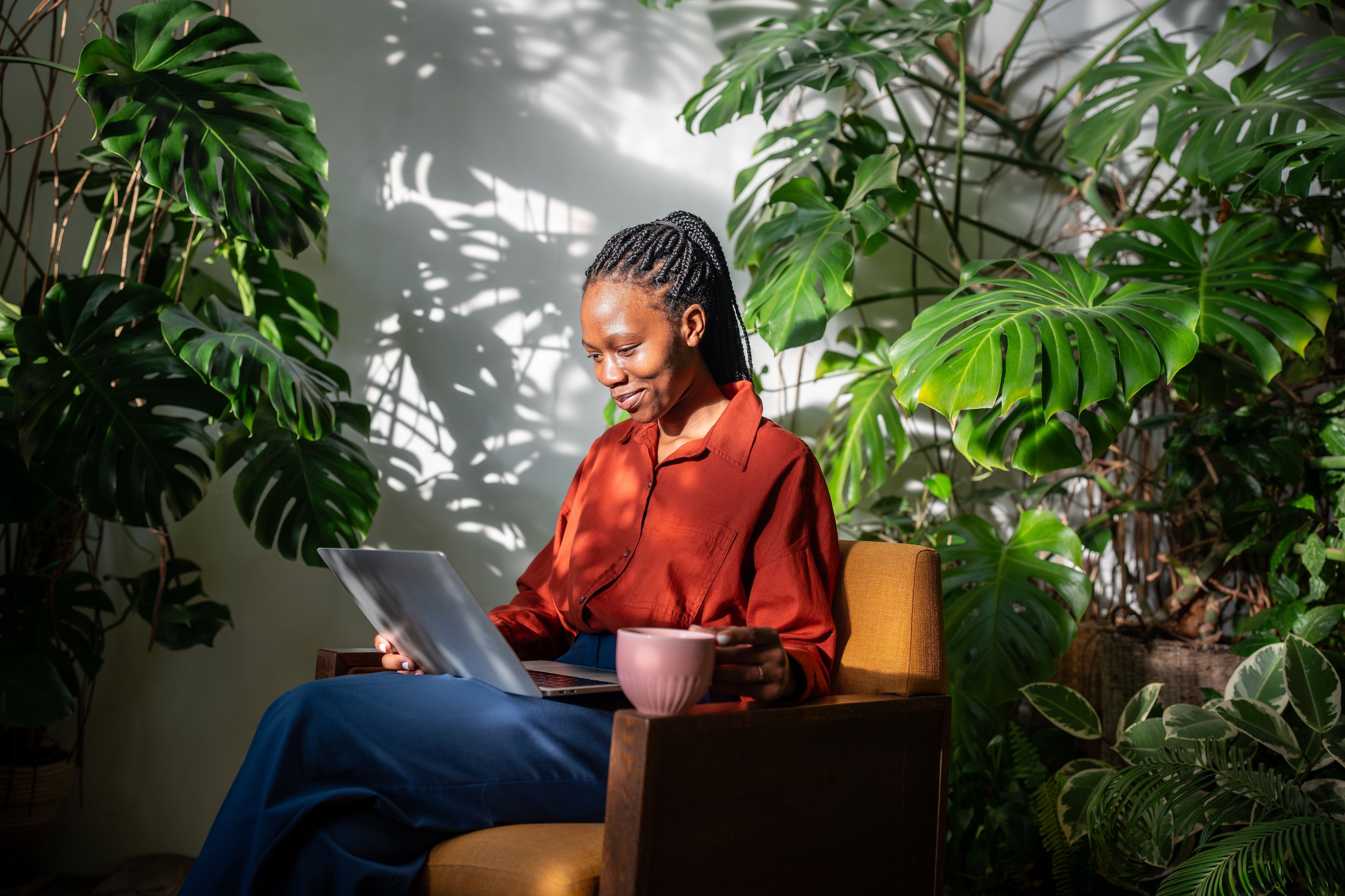 Giggling black woman using laptop. Cheerful laughing african american girl in chair among plants, drinking coffee from mug looking at computer screen, watching video movie series online on internet.