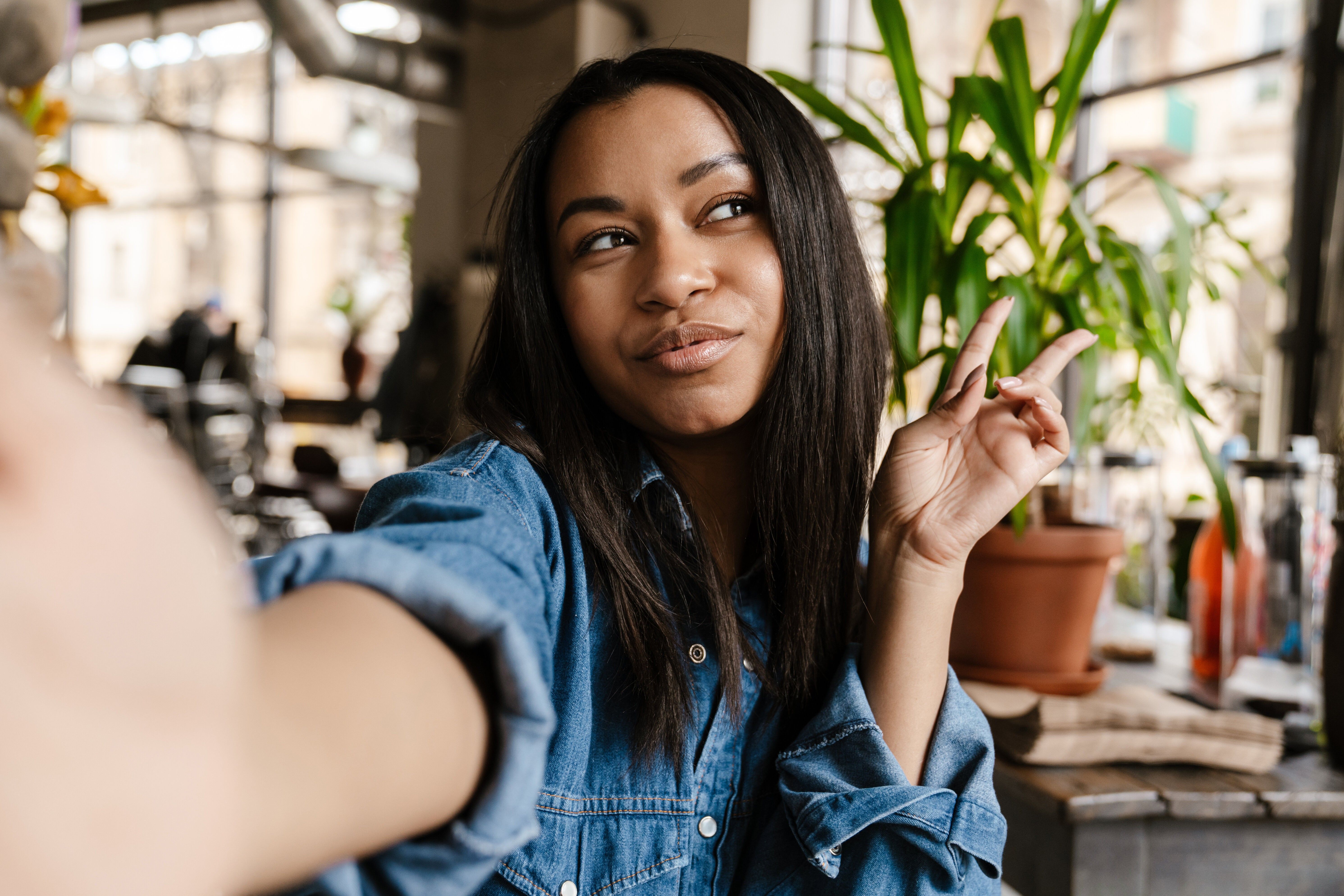 Black smiling woman gesturing peace sign and taking selfie photo while sitting in cafe