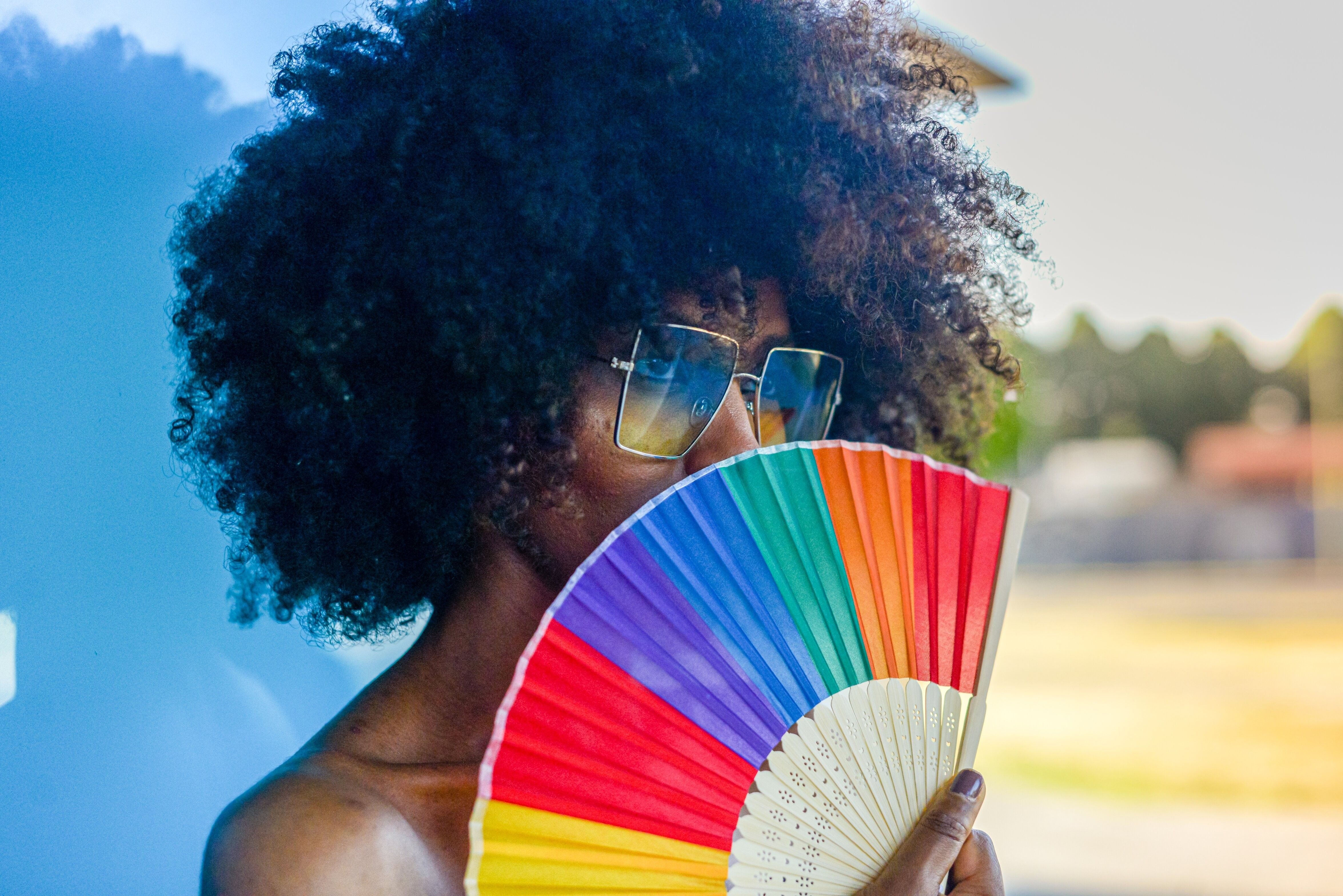 Young Black woman with afro hair and sunglasses cooling off with a colorful rainbow hand fan in summer heat