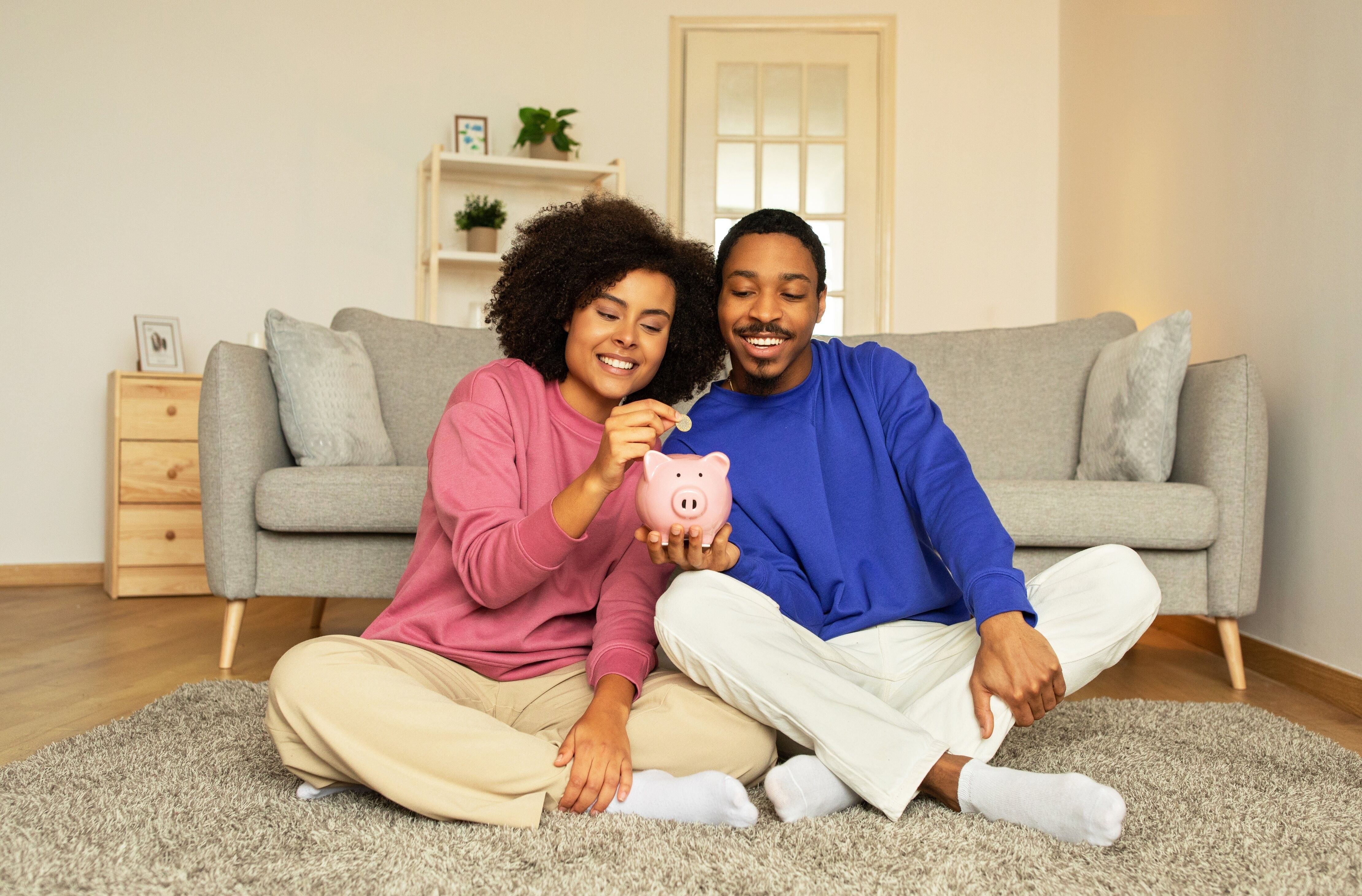 Young African American married couple happily posing with piggy bank, putting money savings sitting together at home. Concept of financial stability, smart money management and future wealth
