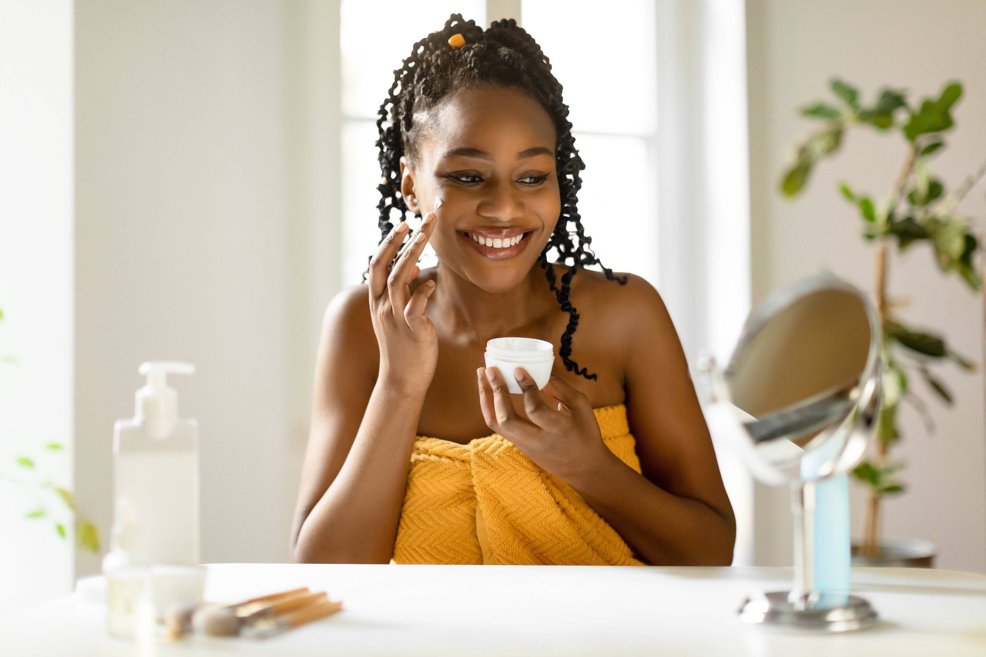 Cheerful black lady wrapped in towel using facial cream after shower, making skin ready for makeup, sitting at table at home, looking at mirror, free space