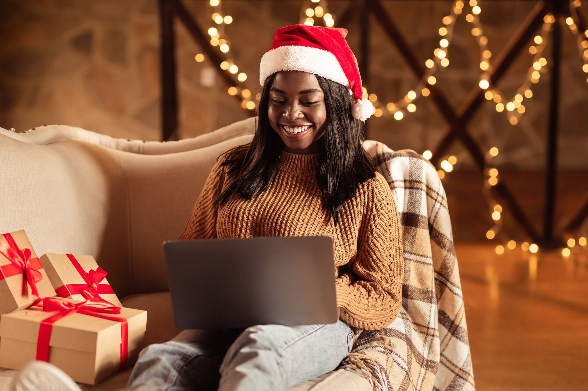 Happy black lady in Santa hat using laptop, ordering Xmas gifts on web, shopping for Christmas presents online from home. African American woman sitting on couch with computer on winter holidays