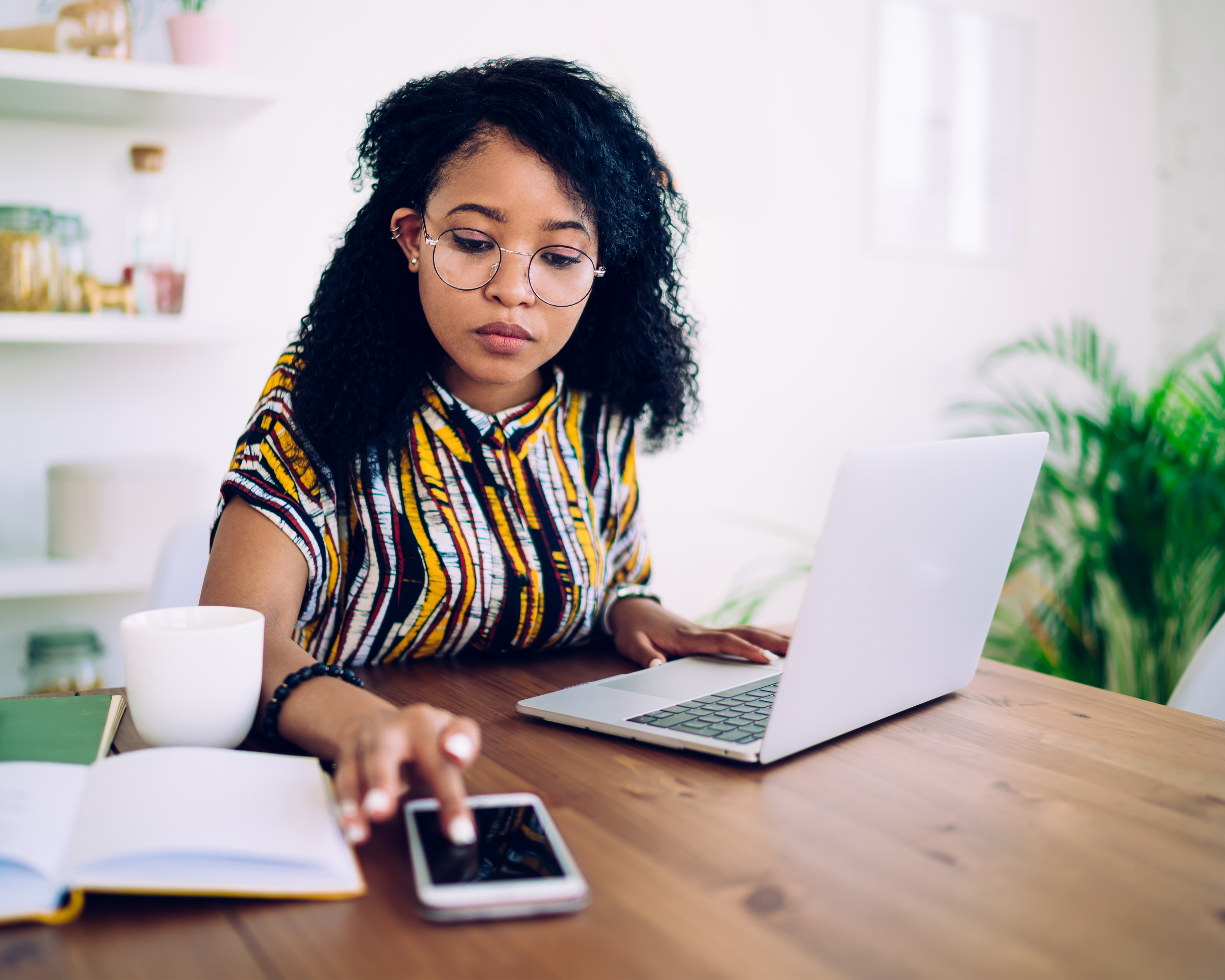 Focused ethnic female freelancer in glasses browsing smartphone and laptop while sitting at table with cup of coffee at home