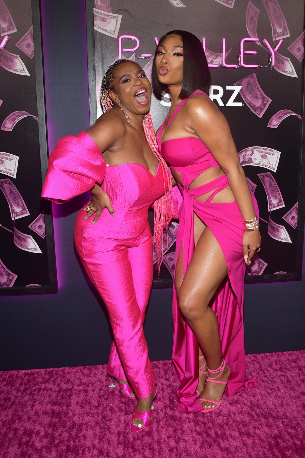Katori Hall stands in a pink, heart-shaped jumpsuit with her hand on her hip, laughing next to Megan Thee Stallion, who is in a pink cutout dress with her lips pursed in a kiss