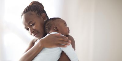 Black mother with brown microbraids and a diamond earring in her ear looks sad as she holds a sleeping infant wearing a blue onesie