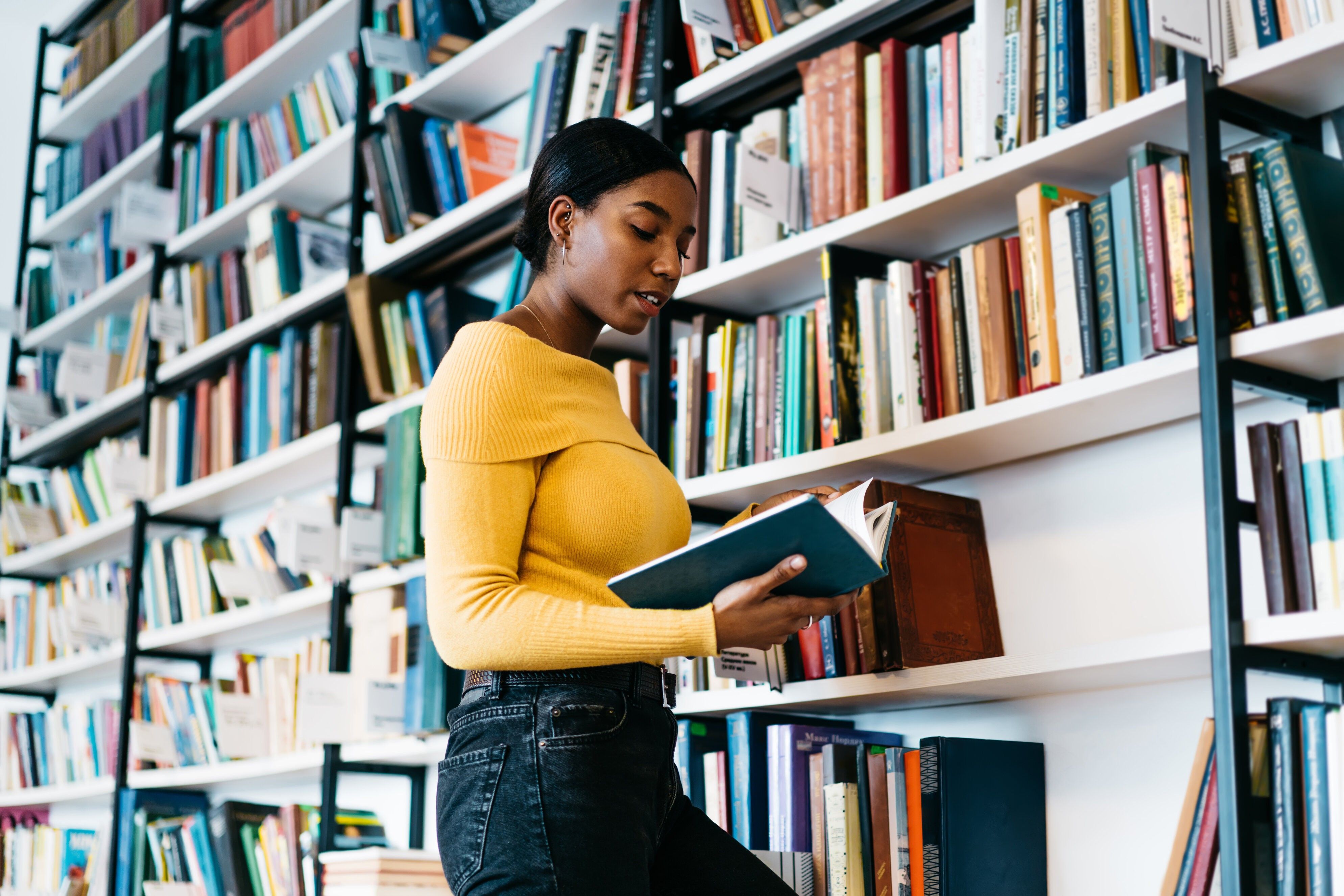 Focused African American lady in casual clothes reading textbook while standing near bookcase and studying in library of contemporary university