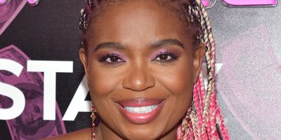 A Black woman, Katori Hall, is seen from the chest up, smiling, in a pink, heart-shaped boostier, and long pink and blonde cornrows