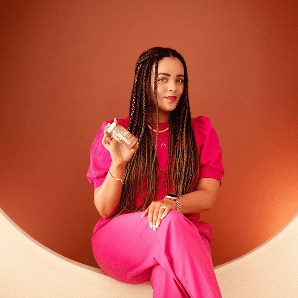 Mela-Vitamins-founder-sitting-in-a-beige-circle-holding-supplements-in-her-hand-crossed-legs