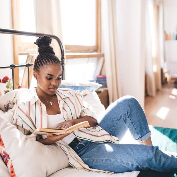 Stylish young Black woman sitting on bed, deeply immersed in reading book, with relaxed body language and casual clothes in sunlit minimalist bedroom