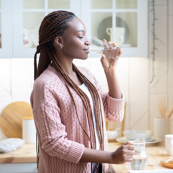 Thirsty African American Woman Drinking Water From Glass In Kitchen At Home, Young Black Female Standing Near Counter And Enjoying Heathy Refreshing Drink, Side View With Copy Space