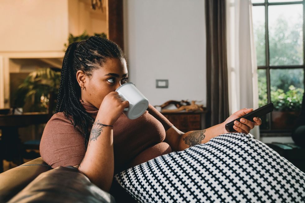 woman-with-braids-drinking-warm-beverage-sitting-on-the-couch-using-the-remote-control