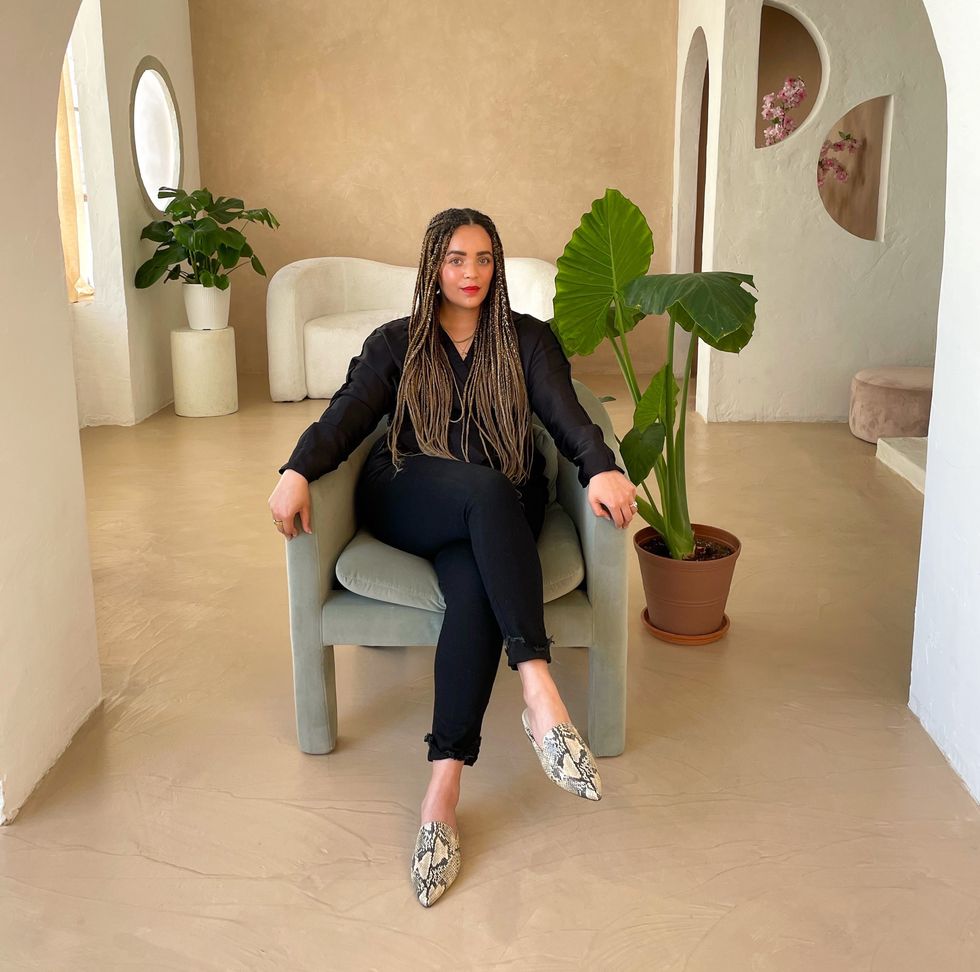 woman-with-braids-sitting-in-a-chair-facing-camera-with-beige-background-green-plant